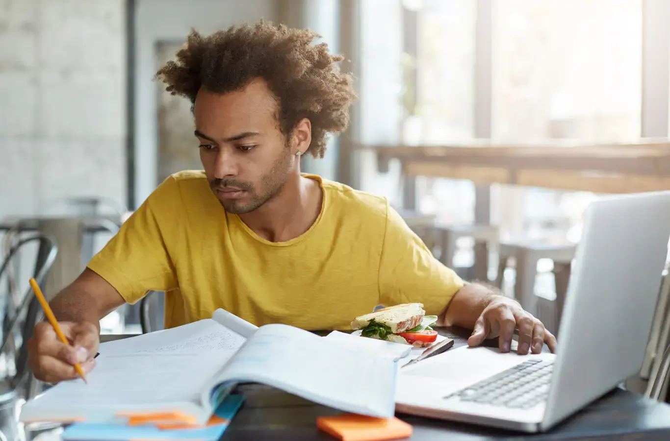 menino escrevendo em um caderno e com o notebook aberto em uma mesa estudando sobre a diferença de isso e isto