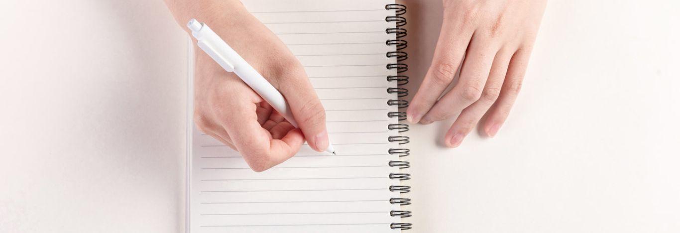 Mão iniciando a escrita de uma resenha em um caderno em branco segurando uma caneta branca.