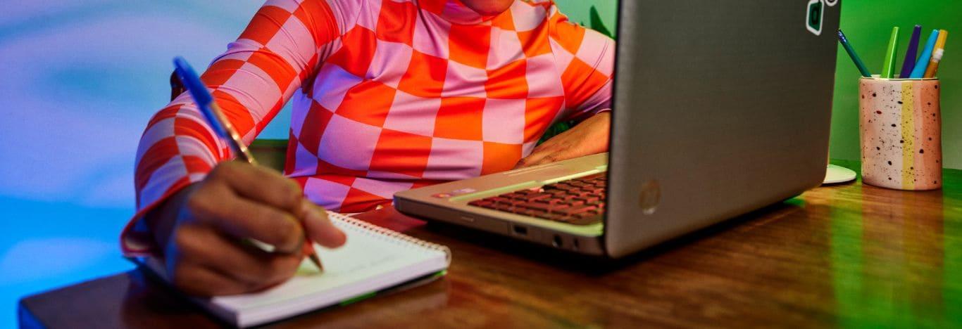 um notebook em cima de uma mesa e uma pessoa resolvendo questões comentadas de naturalismo em um caderno que está do lado do notebook