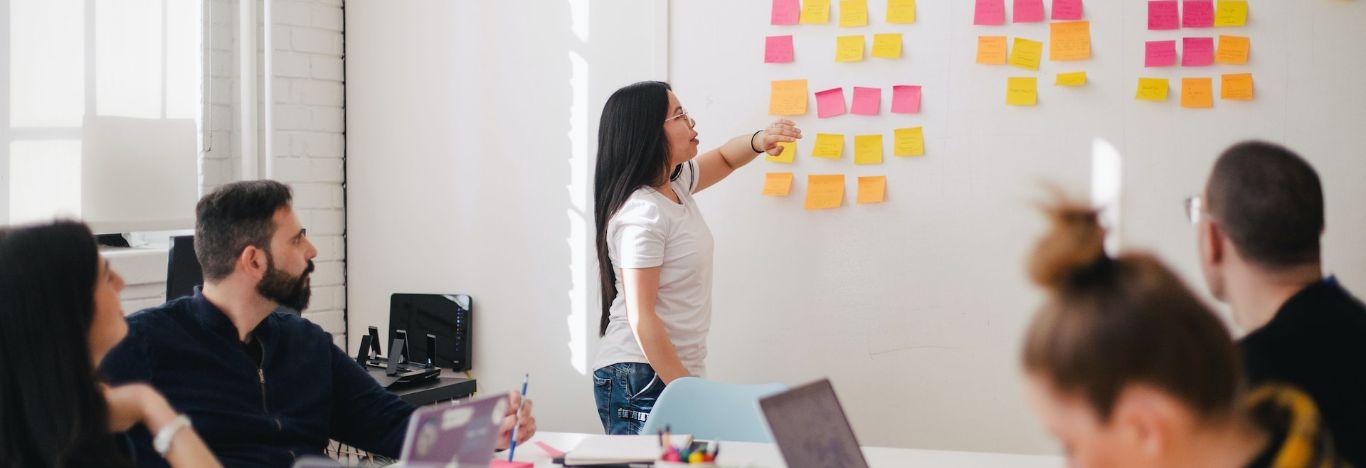 mulher de cabelo longo mostrando uma lousa com alguns post its e pessoas sentadas em volta de uma mesa olhando para ilustrar o contedo sobre pós-graduação em marketing