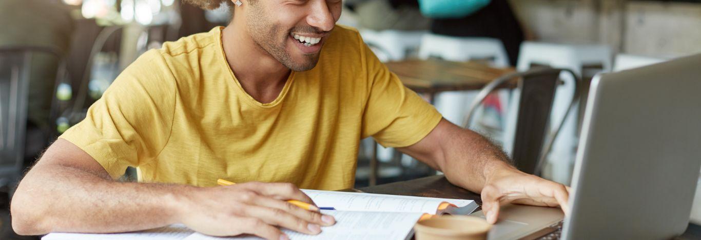 menino sorrindo, lendo um conteúdo sobre a lista de espera do SiSU em um notebook e com a mão em cima de um caderno sob a mesa