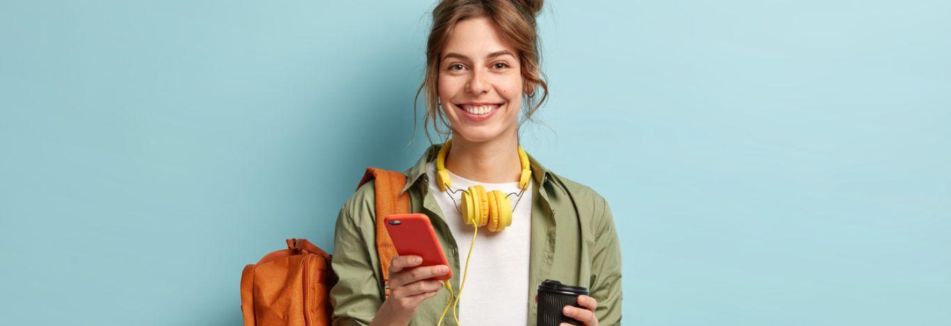 menina de cabelo preso, segurando um celular em uma mão com um conteúdo sobre Enem 2023 aberto, uma xícara em outra mão, com uma mochila no braço, um headset no pescoço e sorrindo para 